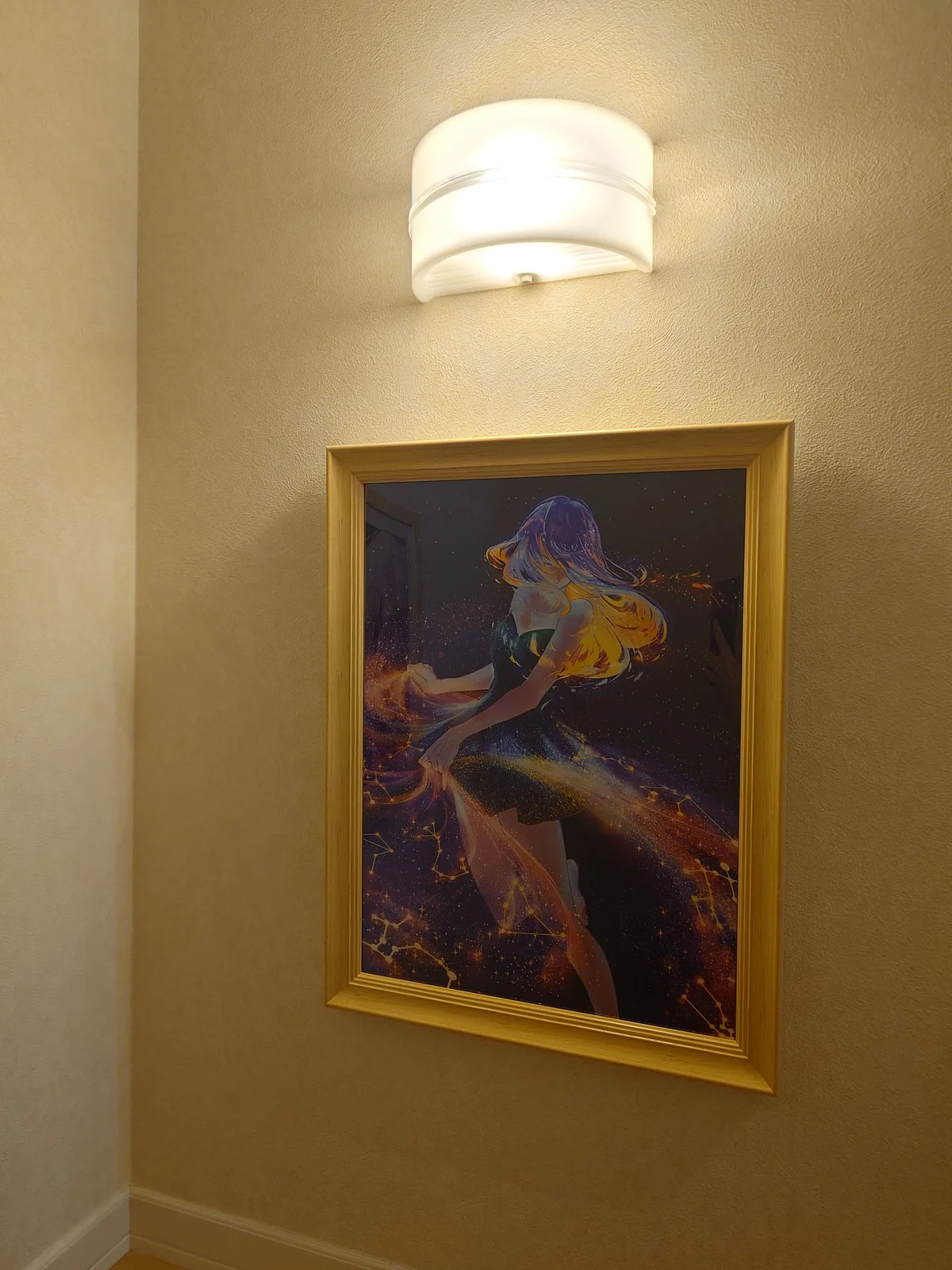 クリーム色の壁に、白い額縁が1点掛かっている（ライトの光で金色っぽく写っている）。中は夜空のような濃い紫と青の背景に星座の線が走り、金色とオレンジ色の髪の人物が布をひるがえしている絵。額縁の真上に乳白色のカバーが付いたブラケットライトが点いていて、その光が上から絵に落ちている